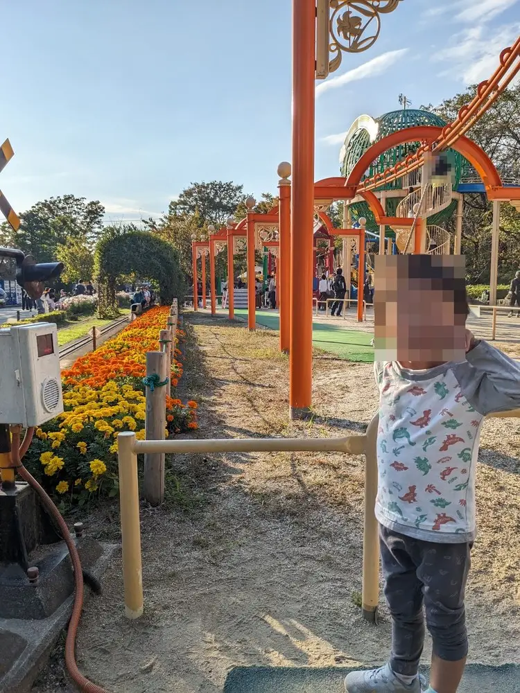 オレンジ色のアーチが続く園路と、その脇に咲き並ぶマリーゴールドの花壇。奥に緑のドーム型遊具